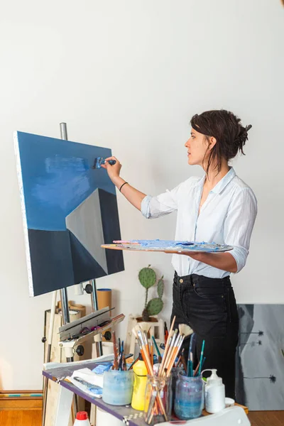 A young smiling painter applies paint to a canvas in her home studio. Other accompanying materials for painting could be seen around it. In some photos, she holds a palette of paints in one hand, while with the other she applies the paint with a pain