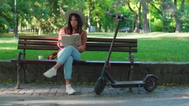 Şapkalı genç ve güzel bir kız kahve içerken bir bankta elinde kitapla oturuyor. Yanında elektrikli bir scooter park ederken, arka planda ağaçlar ağır basıyor..