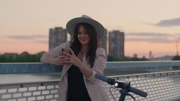 一位面带微笑的年轻女子靠在桥栏上 在电话上打字 一辆电动车停在她旁边 日落和一座现代桥梁的各个部分在背景中占主导地位 — 图库视频影像