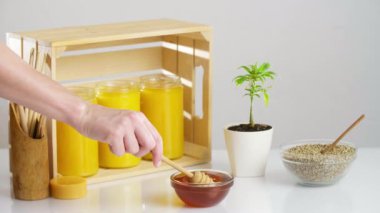 Tahta bir sandığa konmuş üç kavanoz CBD balı. Etrafta marihuana bitkisi, kenevir tohumu dolu bir kase bal için tahta kaşıklar, bir cam kaseye bal damlayan CBD sabun ürünleri var..