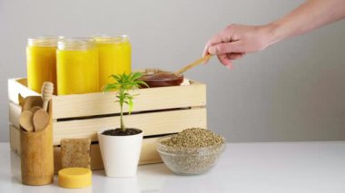 Tahta bir sandığa konmuş üç kavanoz CBD balı. Etrafta marihuana bitkisi, kenevir tohumu dolu bir kase bal için tahta kaşıklar, bir cam kaseye bal damlayan CBD sabun ürünleri var..