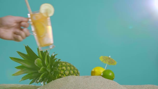 パイナップルやレモンの装飾の横にガラスに氷茶を置く手 背景は砂の半分 青の半分です 夏の雰囲気 — ストック動画