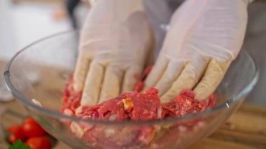 Şefin şeffaf cam bir kâsede köftelerle sebzeleri karıştırırken, iş yüzeyinde sebzeler, pastırma dilimleri, baharatlar ve küçük mutfak aletleri var..
