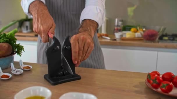 シェフは台所で食事を準備する前にナイフを研ぎます 前景には両手と野菜 スパイス 小さなキッチンボウルが描かれています — ストック動画