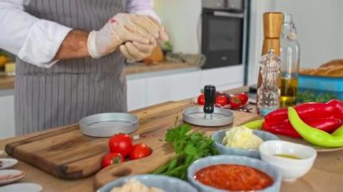 Mutfaktaki ahşap bir tahtanın üzerinde hamburgerler için et şekillendiren bir şefin yakın çekimi. Çeşitli baharatlar, sebzeler ve iş yüzeyinin etrafında küçük mutfak kaseleri var..