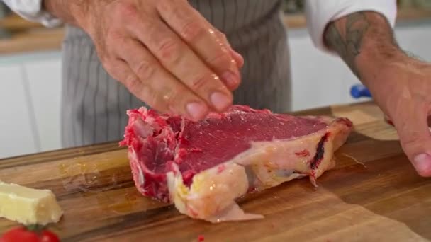 木製のキッチンボード上の肉にオリーブオイルをこすりシェフのクローズアップ 小さなキッチンプレート チェリートマト ナイフなど 作業面の周りに見えます — ストック動画