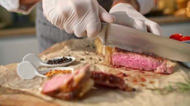 Ahşap bir mutfak tahtasında ızgara bifteği tutup ince parçalara ayıran bir şefin yakın çekim görüntüsü. Yan kaplarda küçük kaseler, baharatlar ve taze ekmekler görülebilir..
