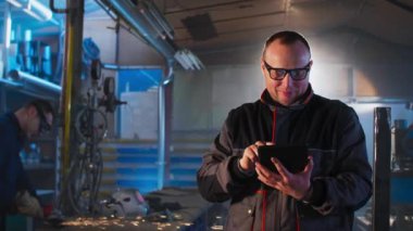 Atölyesinde tableti olan tamirci bir şeyler yazarken, arka planda meslektaşı öğütücü üzerinde çalışıyor..