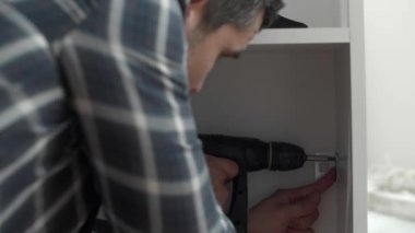 Marangoz vidaları vidalıyor ve mutfak parçalarını elektrikli tornavidayla birleştiriyor. Etrafında iş aletleri ve çalıştığı malzemeler var..