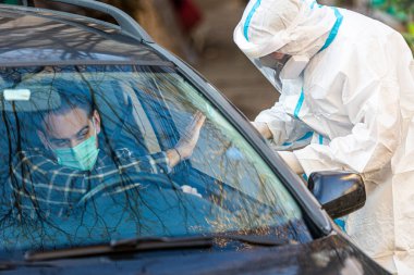 Adam sürüş için COVID 19 testi yapmayı reddediyor, hastadan araba camından burun örneği alıyor, PCR teşhisi koyuyor, PPE 'de elinde test kiti olan bir doktor..