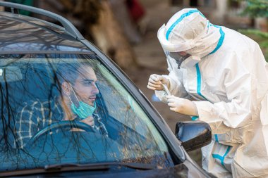 Arabada oturan bir adam, tıbbi çalışanın COVID-19 testi yapmasını bekliyor. Arabanın camından burun temizleme örneği alıyor. Coronavirus için PCR testi yapıyor..