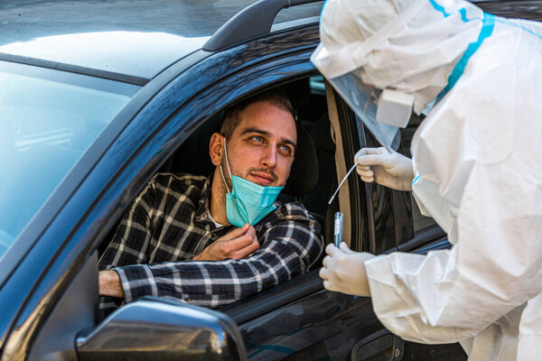 Man sitting in car, waiting for medical worker to perform drive-thru COVID-19 test, taking nasal swab sample through car window, PCR diagnostic for Coronavirus, doctor in PPE holding test kit.