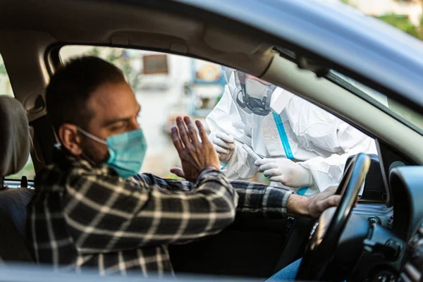 Adam sürüş için COVID 19 testi yapmayı reddediyor, hastadan araba camından burun örneği alıyor, PCR teşhisi koyuyor, PPE 'de elinde test kiti olan bir doktor..