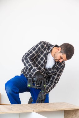 Marangoz vidaları vidalıyor ve mutfak parçalarını elektrikli tornavidayla birleştiriyor. Etrafında iş aletleri ve çalıştığı malzemeler var..