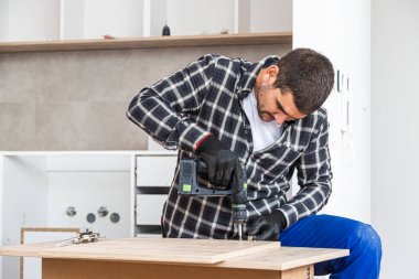 Marangoz vidaları vidalıyor ve mutfak parçalarını elektrikli tornavidayla birleştiriyor. Etrafında iş aletleri ve çalıştığı malzemeler var..