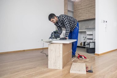 Bir marangoz odanın zemininde çalışma yüzeyinin ahşap bir tahtasını kesiyor. İşini, kendisine bir çalışma masası olarak hizmet eden ahşap bir kutuda yapar..