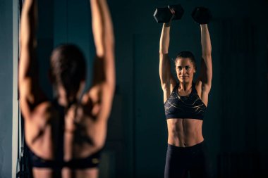 Spor salonundaki aynanın önünde iki dambılla egzersiz yapan genç bir kadın..