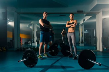 Spor salonunda önlerinde ağırlıklarla poz veren bir çift. Çevrelerinde başka egzersiz ekipmanları var..
