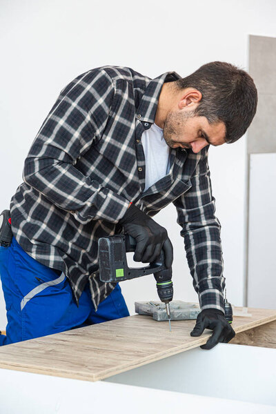 Carpenter screwing screws and assembling parts of kitchen with electric screwdriver. Around him in the room are tools for work, as well as the material he works with.