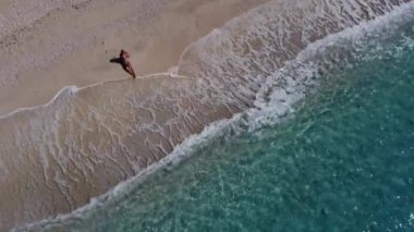 Yüzeysel deniz suyunda kumların üzerinde oturup kumsalın ve yumuşak turkuaz okyanus dalgalarının tadını çıkaran mayolu güzel mutlu kadının hava manzarası. Yaz mevsiminde Lefkada adasındaki Egremni sahilinde tropikal deniz..