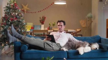 Adam oturma odasındaki kanepede oturuyor Noel süsleriyle, köpeğini okşuyor ve tablet üzerinde çalışıyor..