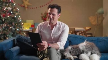 Adam oturma odasındaki kanepede oturuyor Noel süsleriyle, köpeğini okşuyor ve tablet üzerinde çalışıyor..