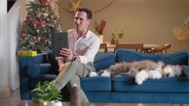 Köpeği yanında otururken adam kanepede oturuyor. Oturma odası Noel ruhuyla dekore edilmiş ve o da tabletiyle birine Noel hediyesi gösteren bir video görüşmesinde..
