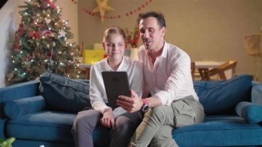 Bir baba ve oğlu oturma odasında kanepede otururken onlar tabletle video görüşmesi yapıp süslü Noel ağaçlarıyla hava atıyorlar. Oda şenlikli bir ruhla dekore edildi. Gelecek Noel 'le birlikte..