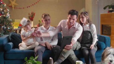 Çocuklarıyla birlikte oturma odasında kanepede oturan bir baba onlar tabletle video görüşmesi yaparken ve süslü Noel ağaçlarıyla hava atarken. Oda şenlikli bir ruhla dekore edildi. Gelecek Noel 'le birlikte..