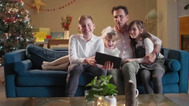 Bir baba, çocuklarının oturma odasında kanepede otururken onlar tabletten bir şey izliyorlar. Oda şenlikli bir ruhla dekore edildi. Gelecek Noel 'le birlikte..