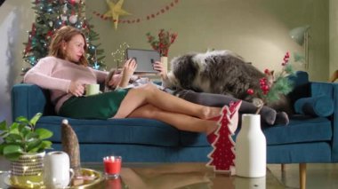 Anne kızı ve bir köpekle kanepede oturuyor. Oda Noel ruhu ile süslenmiştir. Tablette bir şey izlerken gülümsüyorlar ve neşeliler..