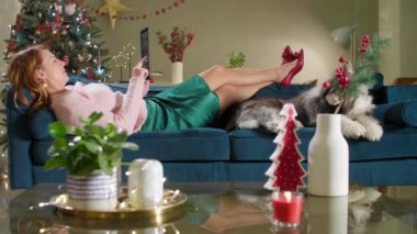 Kadın elinde tabletle kanepede uzanıyor, yanında da evcil bir köpek var. Oturma odası Noel tatili ruhuyla dekore edilmiştir..