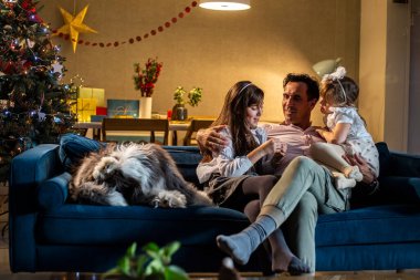 Bir baba oturma odasındaki kanepede iki kızı ve köpeğiyle oturuyor. Evlerinde uzanırken gülümsüyorlar ve neşeliler. Oda Noel ruhuyla süslenmiş..