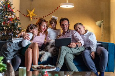Mutlu bir aile oturma odasındaki kanepede oturuyor ve tabletle video görüşmesi yapıyor. Oda Noel ruhuyla süslenmiş. Ayrıca, köpekleri de onlarla birlikte kanepede..