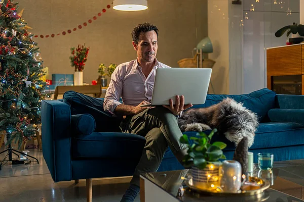 Man working on tablet in living room while his dog is lying next to him. The living room is decorated in the Christmas spirit.