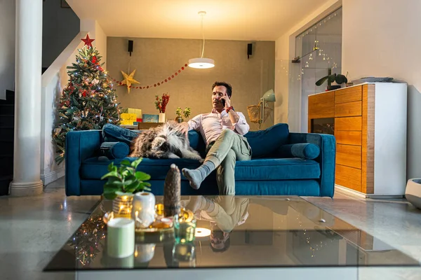 Man is sitting on the couch while talking on a phone. At the same time, he caresses his dog, which is also lying on the couch. The living room is decorated in the spirit of Christmas.