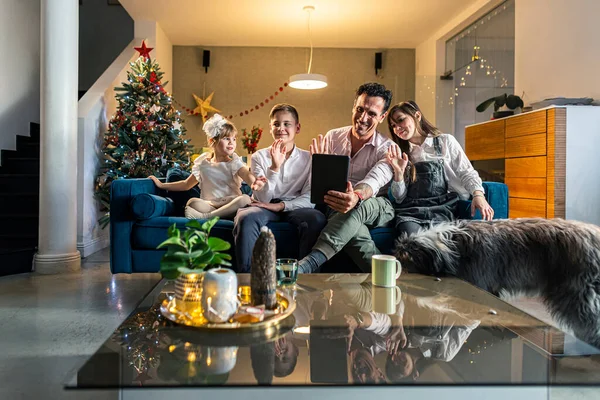 A father with his children sitting in the living room on the couch while they make a video call via tablet and show off their decorated Christmas tree. The room is decorated in a festive spirit, as well as the coming Christmas.