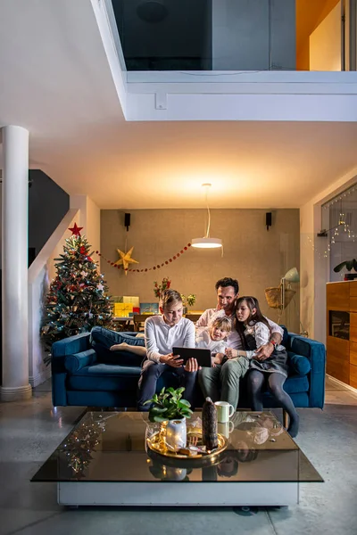 A father sits with his children in the living room on the couch while they watching something on a tablet. The room is decorated in a festive spirit, as well as the coming Christmas.