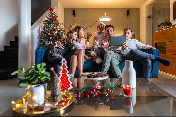 A happy family is sitting on the couch in living room and having a video call via tablet. The room is decorated in the Christmas spirit. Also, their dog is on the couch with them.