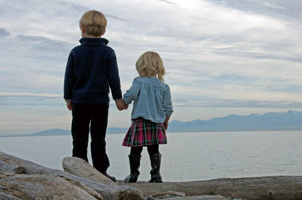Младшие брат и сестра держатся за руки, глядя в море
