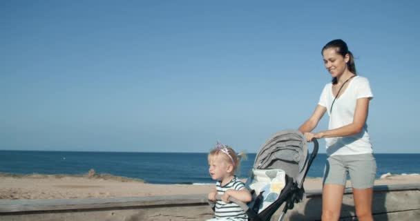 Молодая женщина с дочерью в коляске идет вдоль набережной по морю — стоковое видео