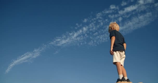 Niño escalando la roca y mirando hacia adelante con el cielo azul nublado como fondo — Vídeo de stock