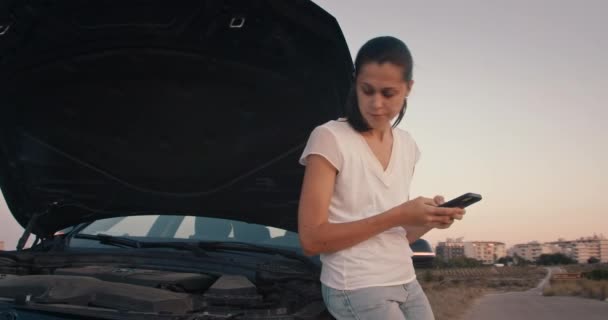 Θλιβερή γυναίκα στέκεται μπροστά στο σπασμένο αυτοκίνητο και χρησιμοποιώντας το smartphone για να βρείτε υπηρεσία — Αρχείο Βίντεο