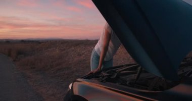 Genç ve çekici bir kadın kırık bir arabanın önünde duruyor ve cep telefonuyla konuşuyor.