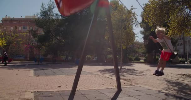 Glückliche hübsche Kinder schaukeln auf Schaukel auf öffentlichem städtischen Spielplatz im Herbstpark — Stockvideo