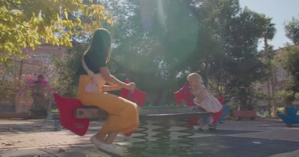 Atraente feliz jovem mãe com bebê menina no playground montando em um balanço — Vídeo de Stock