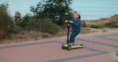 Şapkalı küçük bir çocuk deniz boyunca annesinin peşinden hızla yeşil bir scooter sürüyor.