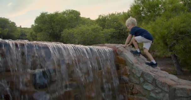 Lindo chico rubio sube a la montaña cerca del agua que cae por la noche en el parque — Vídeo de stock