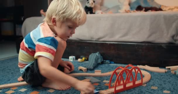 Bonito menino feliz jogando brinquedo de madeira estrada ferroviária na sala de estar à noite — Vídeo de Stock