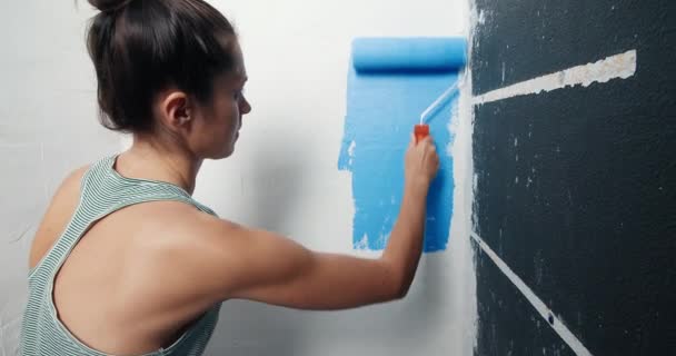 Vista posteriore di attraente giovane donna pittura parete bianca su blu con rullo — Video Stock